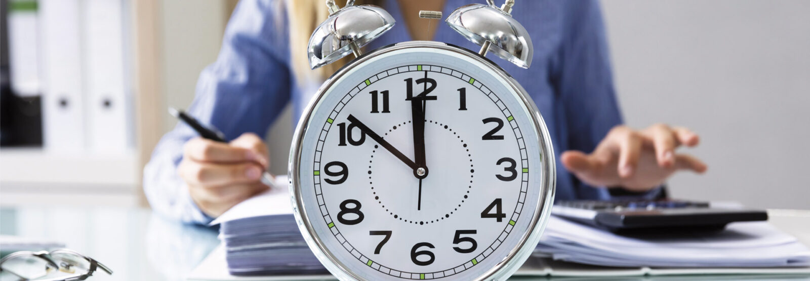 Eine Frau sitzt am Schreibtisch und arbeitet, vor ihr steht eine Uhr, die 10 Minuten vor 12 anzeigt