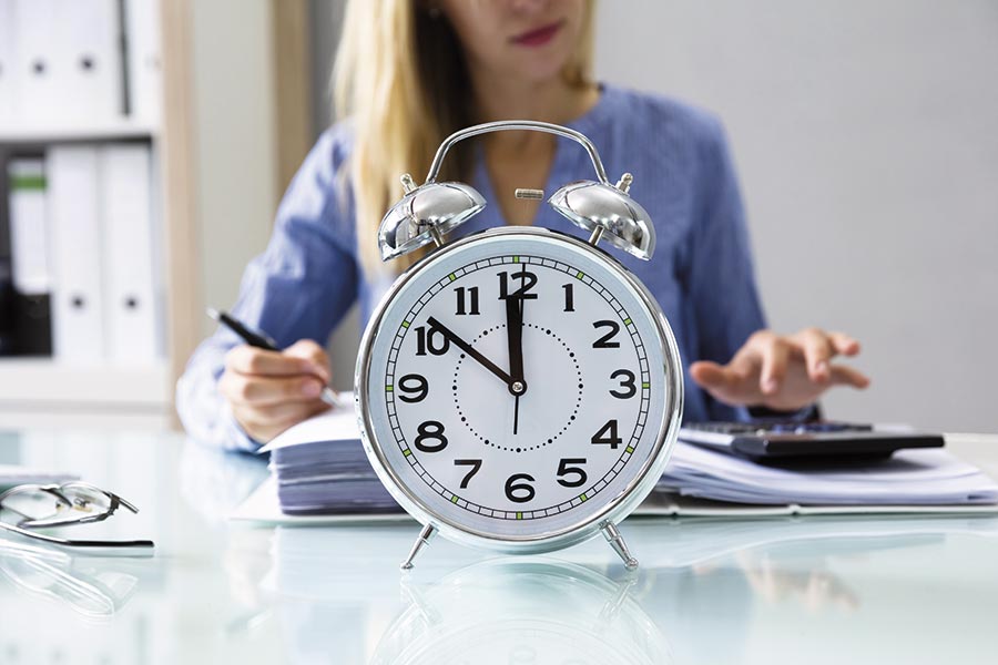 Eine Frau sitzt am Schreibtisch und arbeitet, vor ihr steht eine Uhr, die 10 Minuten vor 12 anzeigt