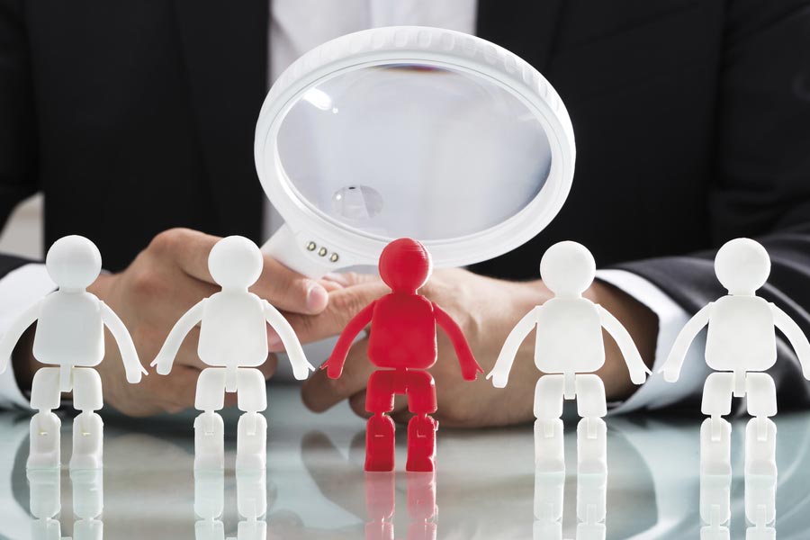 A man in a suit looks through a magnifying glass at a red figure standing between four white figures.