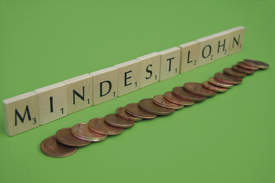 The picture shows the lettering "Minimum wage" made of Scrabble letters on a green background. Cent coins lie in a row in front of it.
