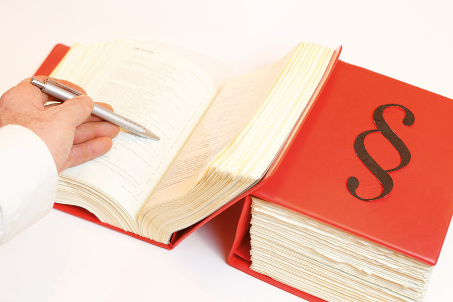 Hand mit Stift geht den Text in einem Buch durch, auf dem ein Paragraphenzeichen abgebildet ist.