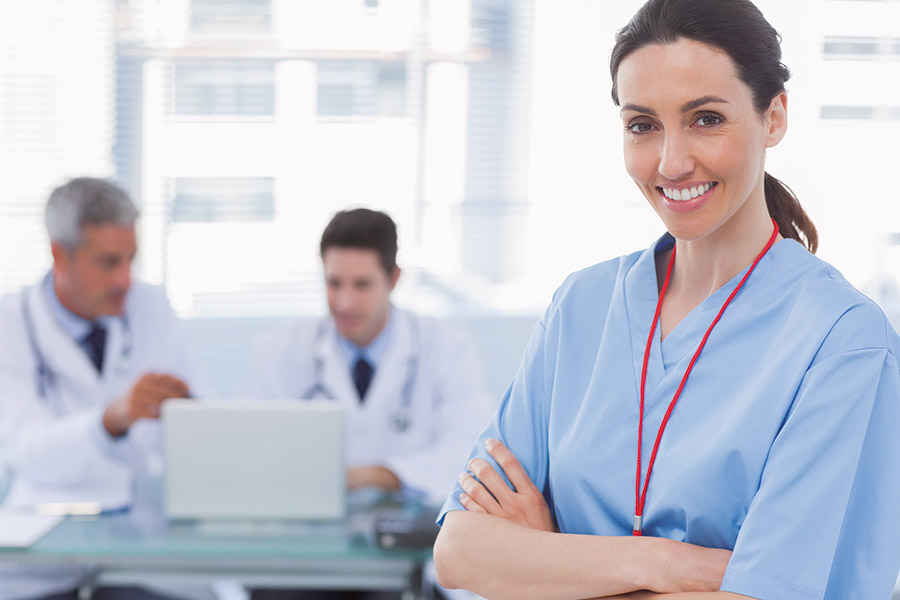 Mediziner arbeiten an einem Laptop während im Vordergrund eine Krankenschwestern die Arme verschränkt hält und lächelt