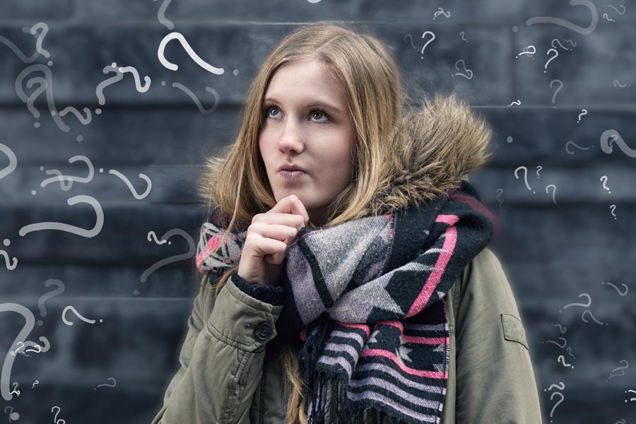 Eine junge Frau steht in einer nachdenklichen Pose vor einer Tafel auf der eine Vielzahl von Fragezeichen in zu erkennen sind.