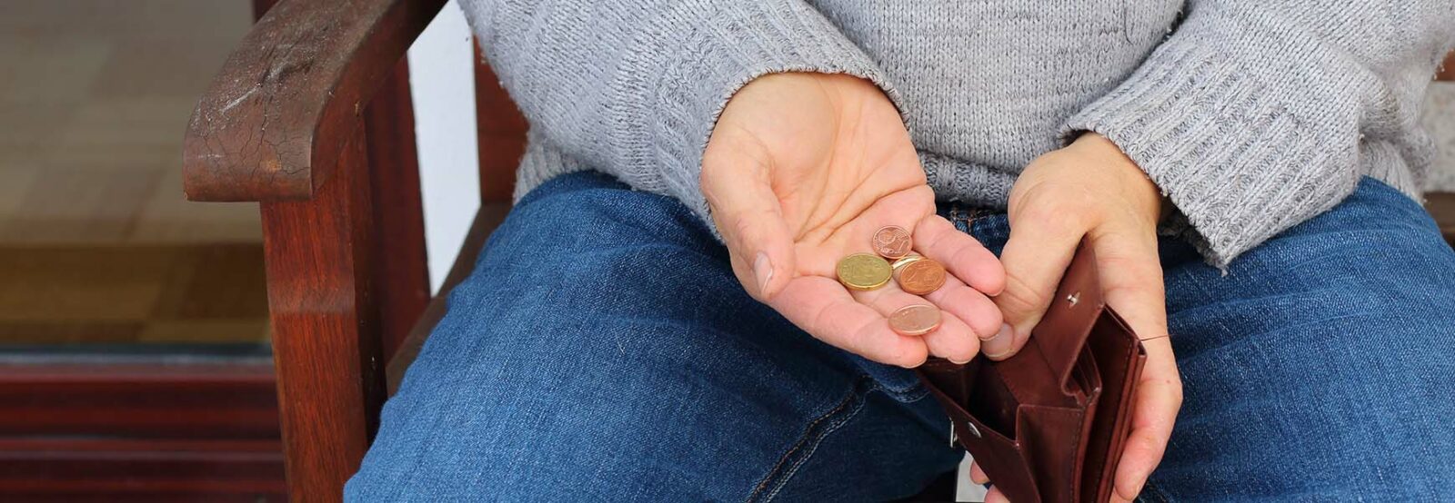 Nahaufnahme von einem Mann, der auf einer Holzbank sitzt, mit dem Fokus auf seinen Händen. In der einen Hand hält er einen offenen Geldbeutel, in der offenen Handfläche der anderen Hand ein paar Münzen.