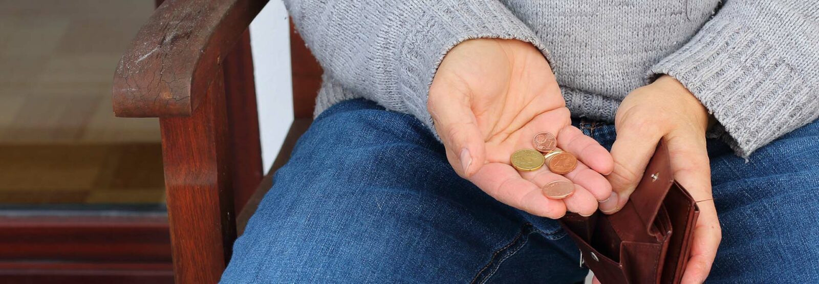 Nahaufnahme von einem Mann, der auf einer Holzbank sitzt, mit dem Fokus auf seinen Händen. In der einen Hand hält er einen offenen Geldbeutel, in der offenen Handfläche der anderen Hand ein paar Münzen.