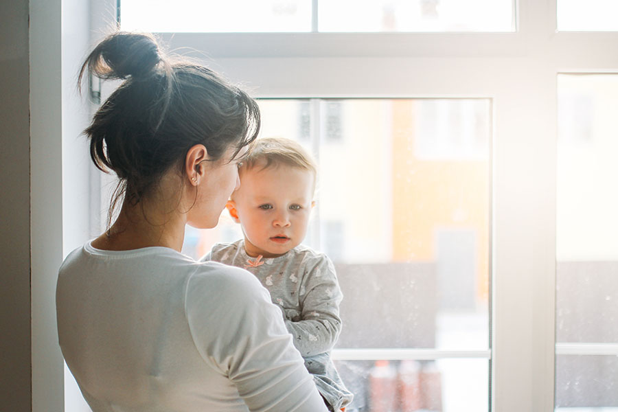 Das Bild zeigt eine junge Mutter von schräg hinten, die Ihr Baby auf dem Arm hält während sie aus dem Fenster schaut.