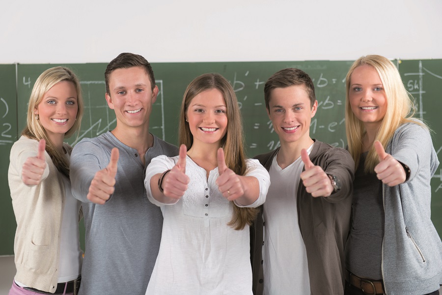 Fünf lächelnde Jugendliche stehen in einem Klassenraum vor der Tafel und zeigen "Daumen hoch".