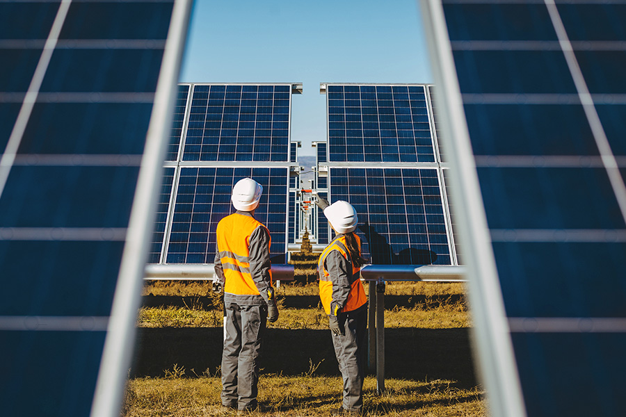 Das Bild zeigt eine Frau und einen Mann mit Arbeitskleidung von hinten in einem Solarpark stehend. Beide schauen in Richtung der Anlage, auf die die Frau deutet.