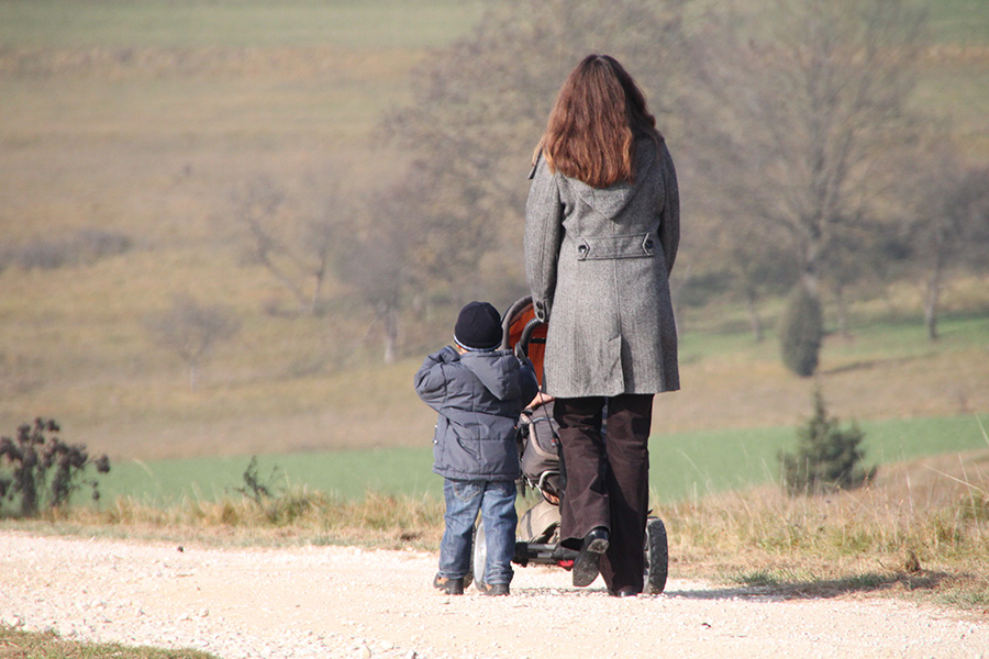 Eine Frau geht mit einem Kleinkind und einem Kinderwagen auf einem Feldweg spazieren