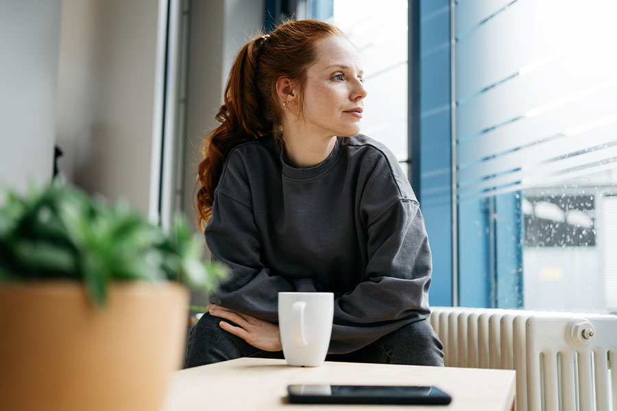 A young woman sits at a table by the window and looks out, lost in thought. There is a cup, a cell phone and a potted plant on the table.