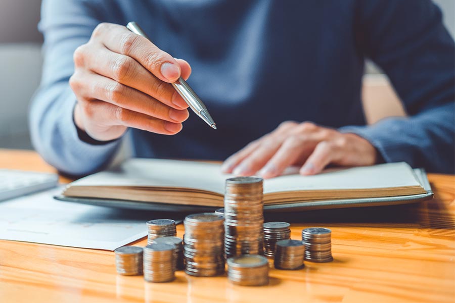 Close-up of a man sitting in front of a notebook and pointing with a pen at stacks of coins of different heights. The focus is on the hand with the pen.