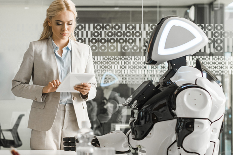 Junge Frau steht mit einem Laptop in der Hand in einem futuristisch anmutenden Büro, in dem ein Roboter auf einem Stuhl sitzt.