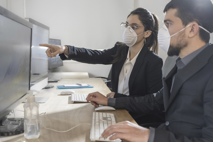 Das Bild zeigt, eine Frau und einen Mann mit Mund-Nasenschutz, die in einem Büro am Schreibtisch sitzen. Die Frau zeigt dem Mann etwas auf dem Bildschirm des Computers.