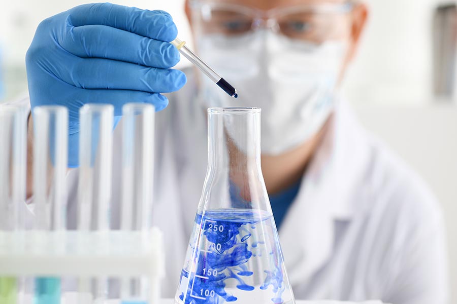 Chemiker mit Schutzbrille, Mundschutz und Gummihandschuhen tropft mit einer Pipette eine blaue Flüssigkeit in einen Erlenmeyerkolben, der mit einer klaren Flüssigkeit gefüllt ist.