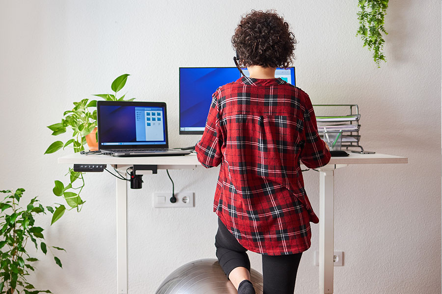 Shot from behind: young woman working standing at her height-adjustable desk, resting her left leg on an exercise ball.