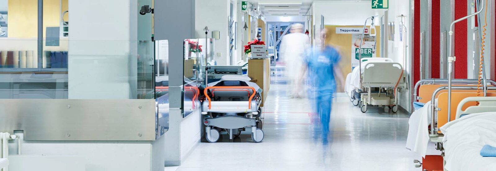 Corridor in a hospital with beds and equipment on the sides. A person from the nursing staff can be seen out of focus in the centre.