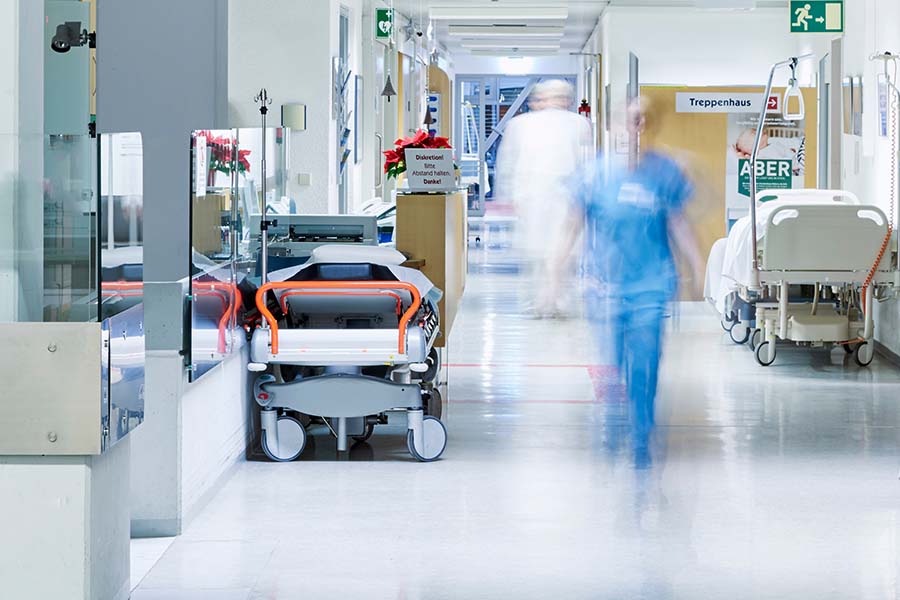 Corridor in a hospital with beds and equipment on the sides. A person from the nursing staff can be seen out of focus in the centre.