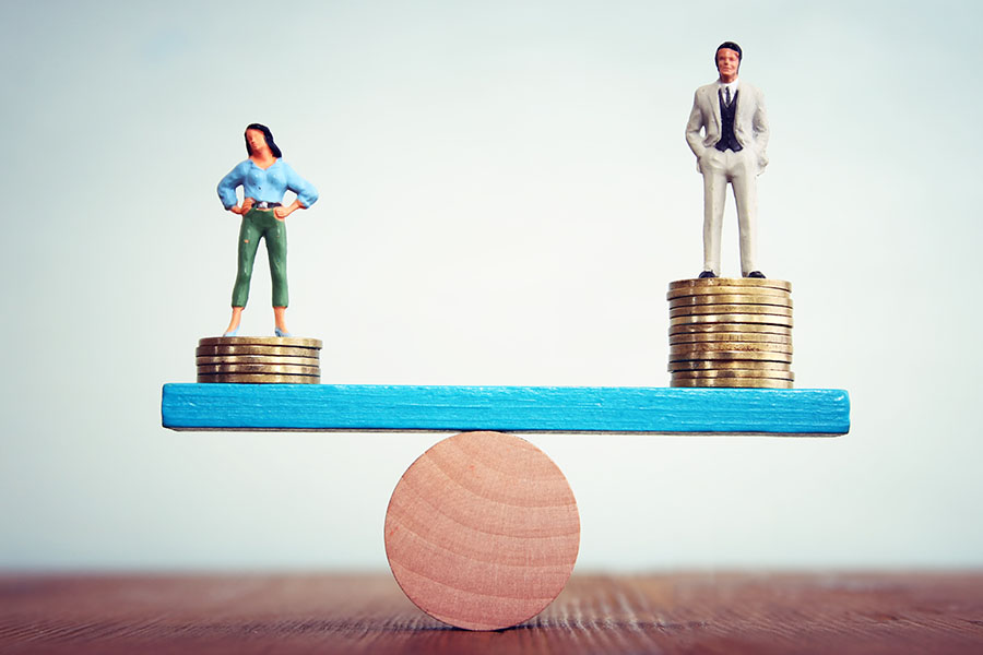 A seesaw with a miniature figure of a woman on one side and a miniature figure of a man on the other. Both are standing on a stack of coins. The woman's stack is smaller, but the seesaw is still balanced.