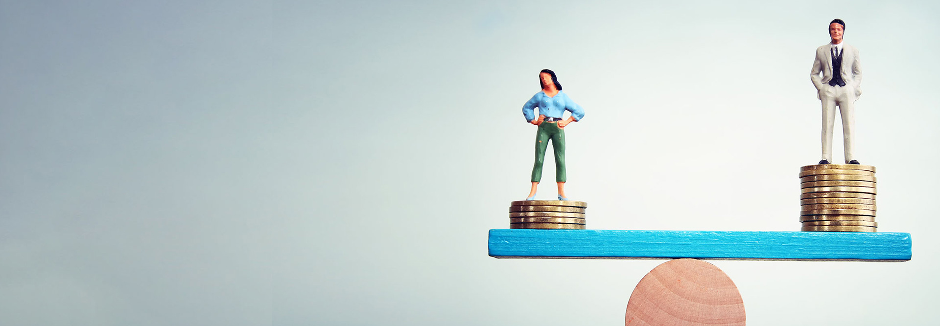 A seesaw with a miniature figure of a woman on one side and a miniature figure of a man on the other. Both are standing on a stack of coins. The woman