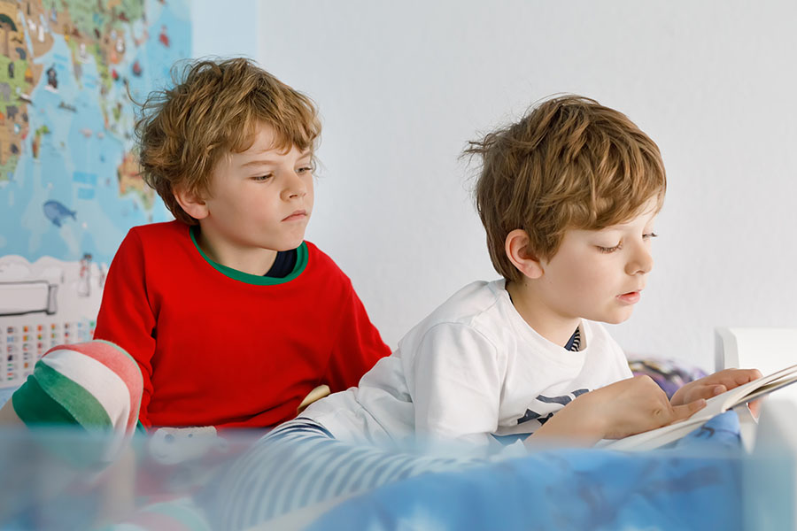 Zwei Jungs in Schlafanzügen sitzen im Bett. Der eine sitzt dem anderen abgewandt und liest ein Buch. Der andere schaut ihm mit hängenden Mundwinkeln über die Schulter.