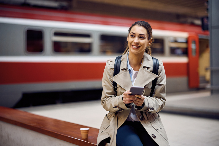 Eine junge Frau mit dunklen Haaren sitzt mit ihrem Smartphone in der Hand auf einer Bank in einem Bahnhof. Hinter ihr ist ein roter Zugwaggon mit geöffneter Türe zu erkennen.