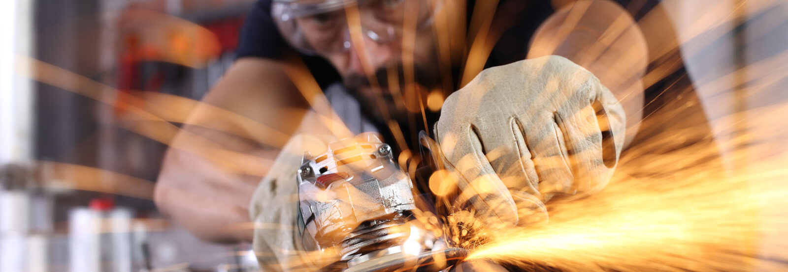 Nahaufnahme von einem Mann, der mit Schutzbrille und Schutzhandschuhen ein Metallwerkstück schleift, das in einem Schraubstock eingespannt ist. Es fliegen orangene Funken.