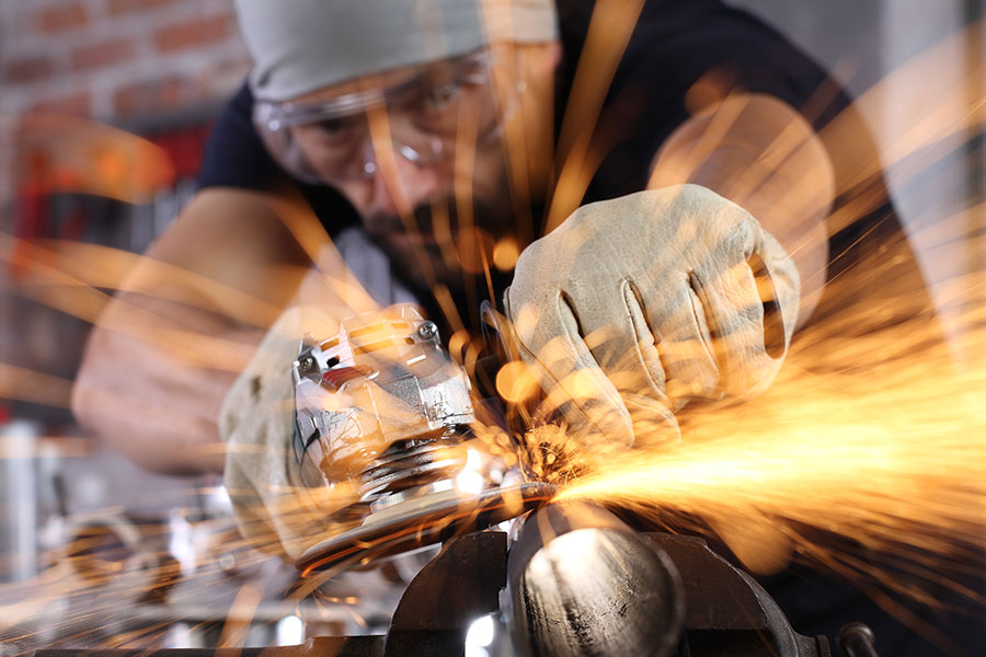 Nahaufnahme von einem Mann, der mit Schutzbrille und Schutzhandschuhen ein Metallwerkstück schleift, das in einem Schraubstock eingespannt ist. Es fliegen orangene Funken.