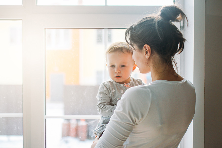 Eine dunkelhaarige Frau hält ein kleines vor einem sonnendurchfluteten Fenster ein Kind auf dem Arm.