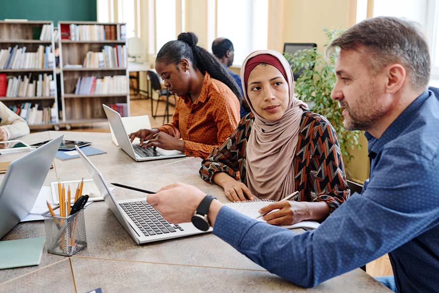 A teacher explains something to an female adult Muslim student wearing a hijab on the laptop they are both sitting in front of.