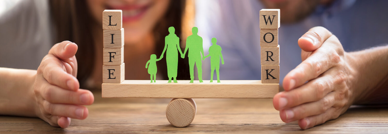 A man and woman hold their hands protectively around a seesaw, balancing on it. On the left is the word "Life" and on the right the word "Work." In the center is a silhouette of a family.