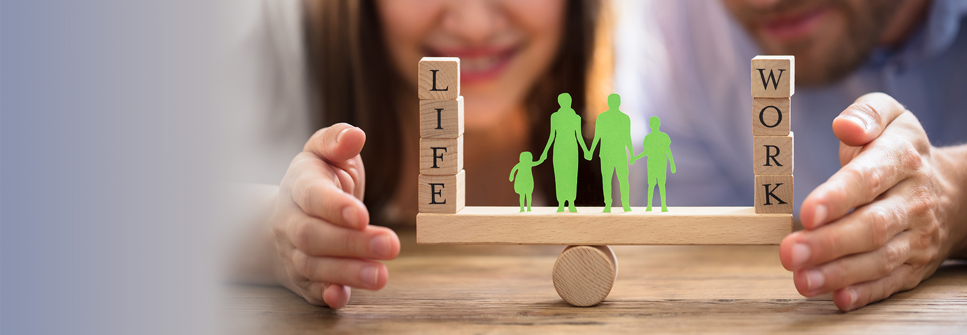 Mann und Frau halten schützend die Hände um eine Wippe in Balance. Auf dieser steht links das Wort „Life“ und rechts das Wort „Work, in der Mitte befindet sich der Scherenschnitt einer Familie.