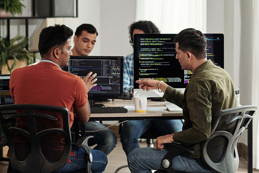 Vier junge Softwareentwickler sitzen an ihren Rechnern und diskutieren. Auf den Monitoren sind Programmiercodes erkennbar.