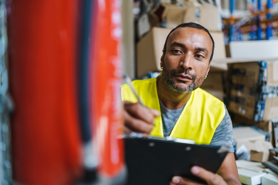 A worker with an Arab appearance checks a fire extinguisher in a warehouse and takes notes.