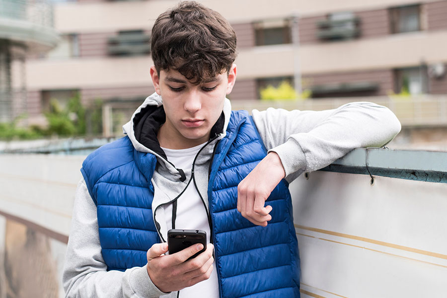 Ein Jugendlicher steht vor einem Wohnblock und schaut auf sein Handy, das er in der Hand hält.