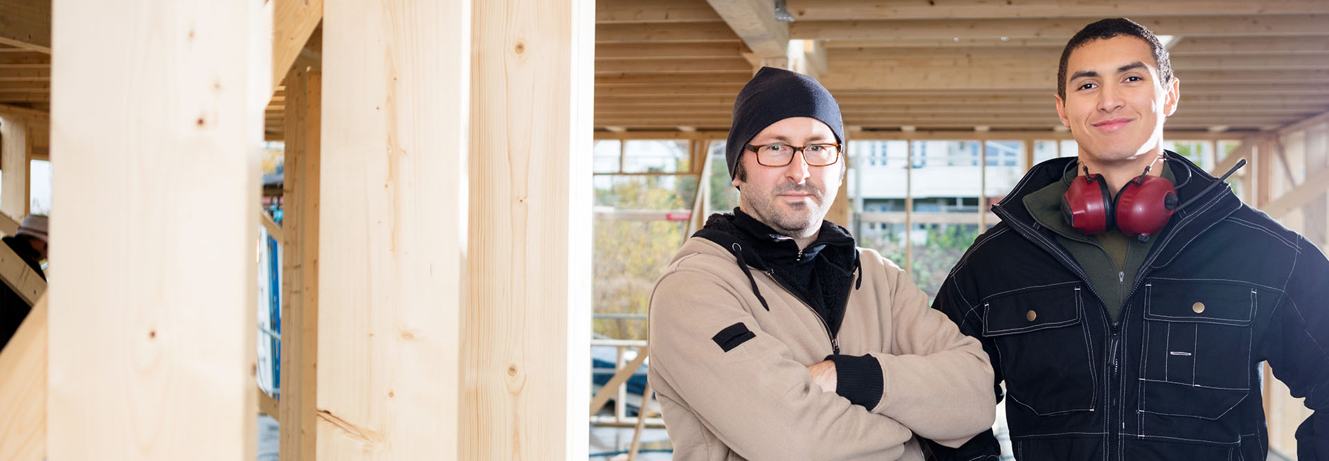 Portrait von zwei Zimmerern, die selbstbewusst auf ihrer Baustelle stehen und zum Betrachter blicken.