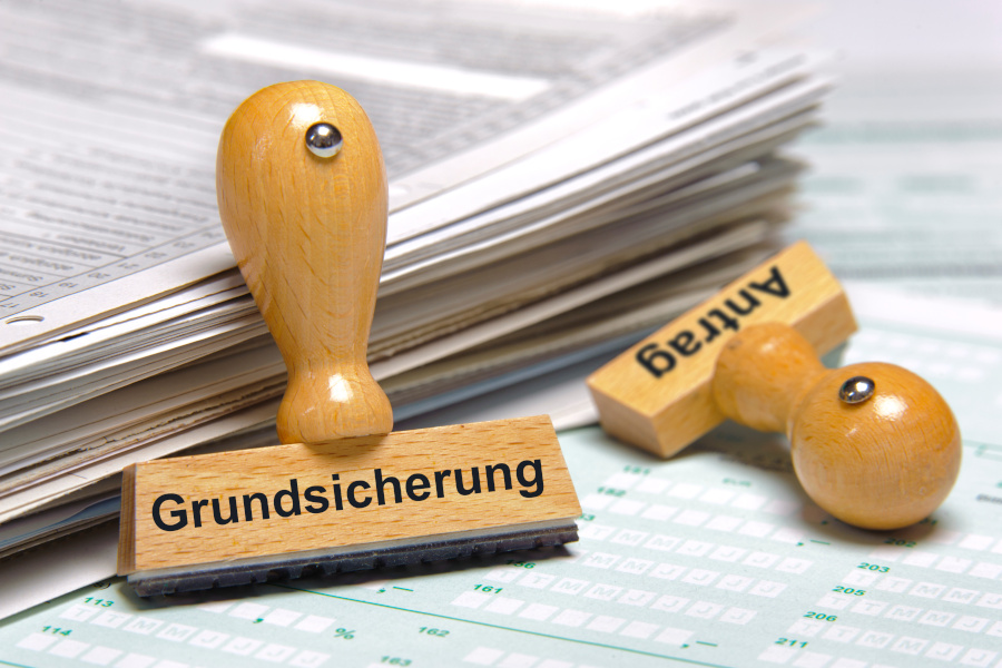 Zwei Stempel liegen vor einem Stapel mit Unterlagen. Einer trägt die Aufschrift „Grundsicherung“ einer das Wort „Antrag“.