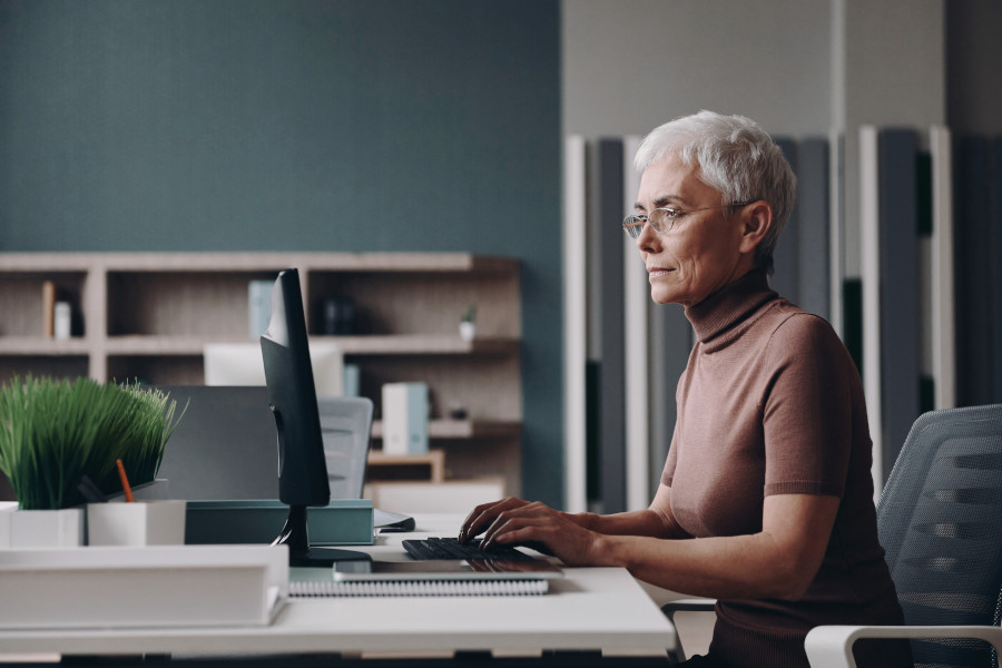 Geschmackvoll gekleidete ältere Frau mit Brille und grauen Haaren arbeitet im Büro an einem Computer.