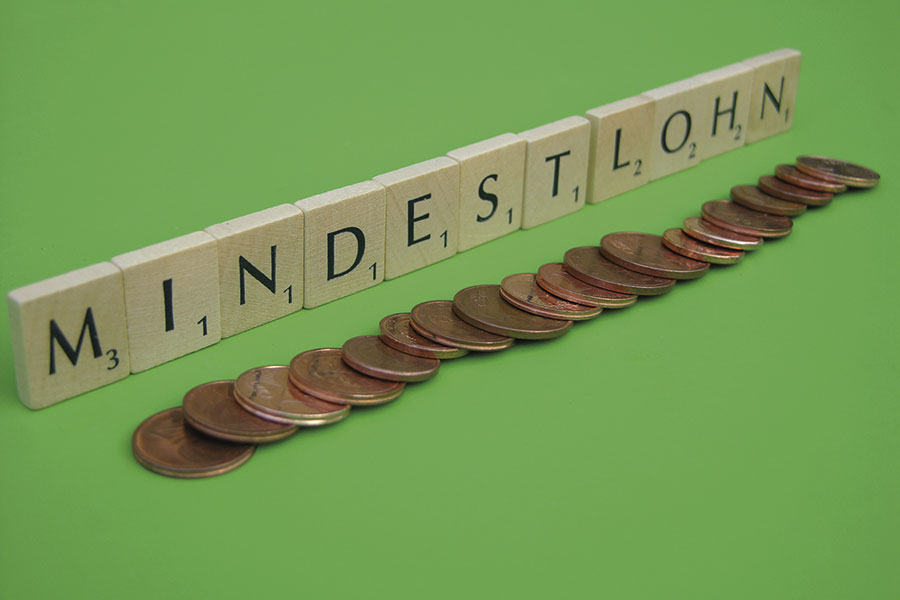 Das Bild zeigt den Schriftzug „Mindestlohn“ aus Scrabble-Buchstaben auf einem grünen Hintergrund. Davor liegen Cent-Münzen in einer Reihe.