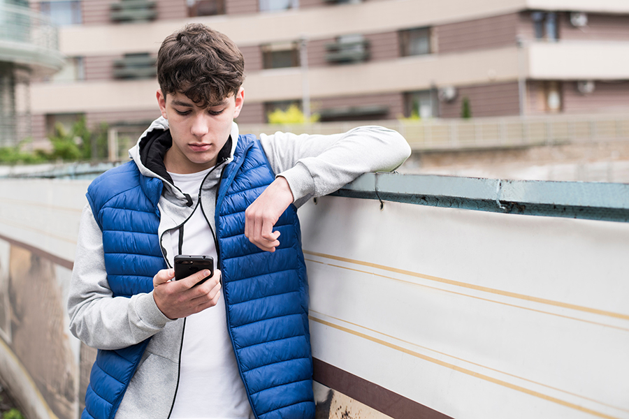 Ein Jugendlicher steht vor einem Wohnblock und schaut auf sein Handy, das er in der Hand hält.