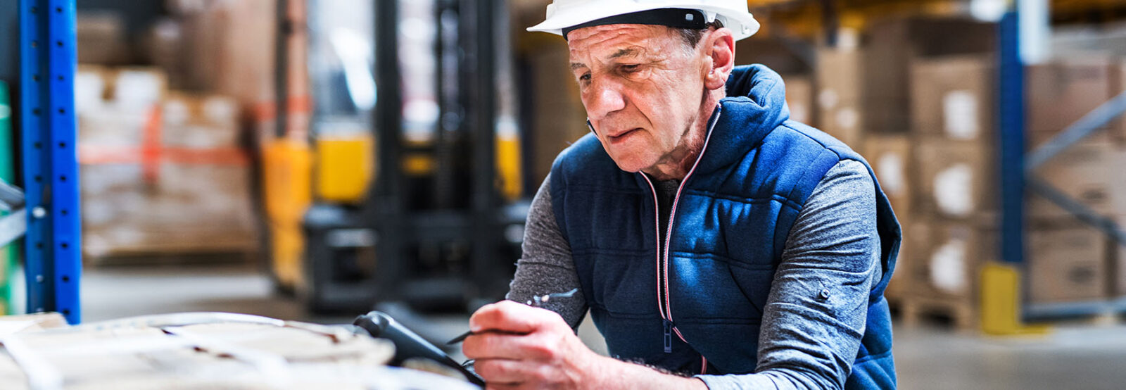 Älterer Mann mit Schutzhelm bei der Arbeit an einem Tablet in einem Lagerhaus.