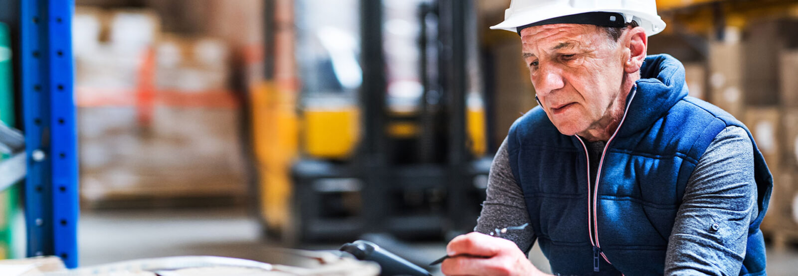 Älterer Mann mit Schutzhelm bei der Arbeit an einem Tablet in einem Lagerhaus.