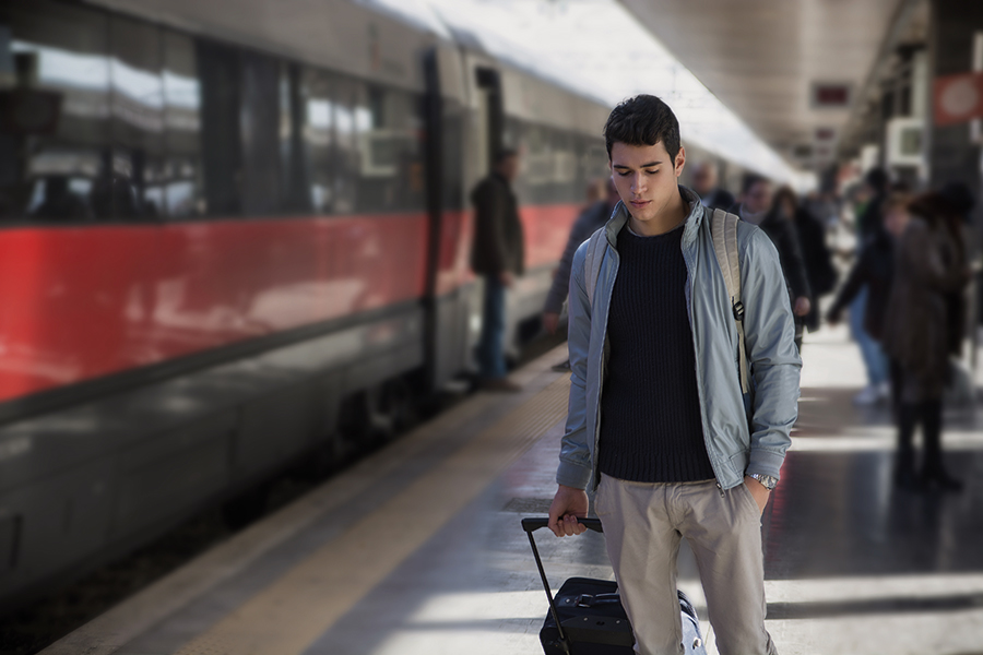 Junger dunkelhaariger Mann läuft mit einem Rollkoffer an einem am Bahnsteig stehenden Zug entlang. Er hat den Blick nach unten gerichtet.