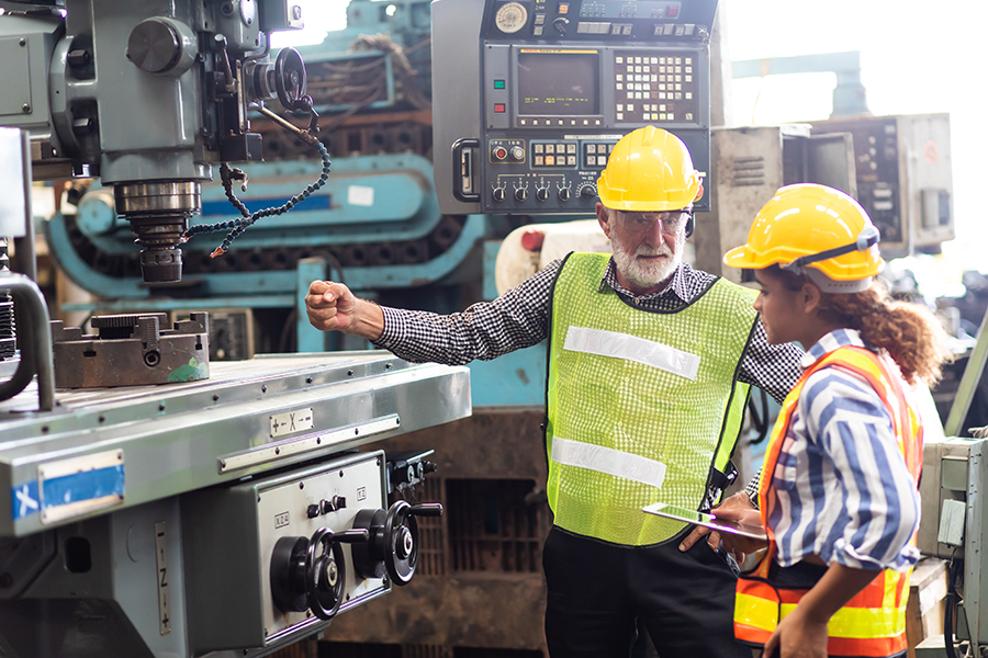 Ein älterer grauhaariger Arbeiter erklärt einer jungen Arbeiterin die Funktion einer Metalldrehmaschine.