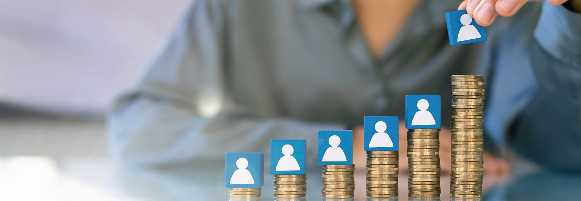 Hand greift Würfel mit Figur, der auf aufsteigenden Stapeln Geldmünzen steht.