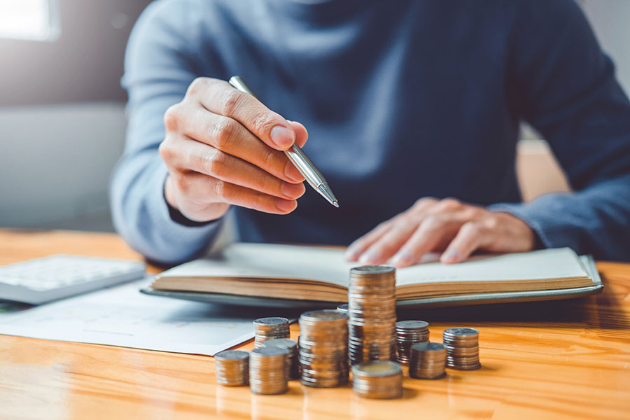 Nahaufnahme eines Mannes, der vor einem Notizbuch sitzt und mit dem Kugelschreiber auf verschieden hohe Münzstapel zeigt. Der Fokus liegt auf der Hand mit dem Kugelschreiber.