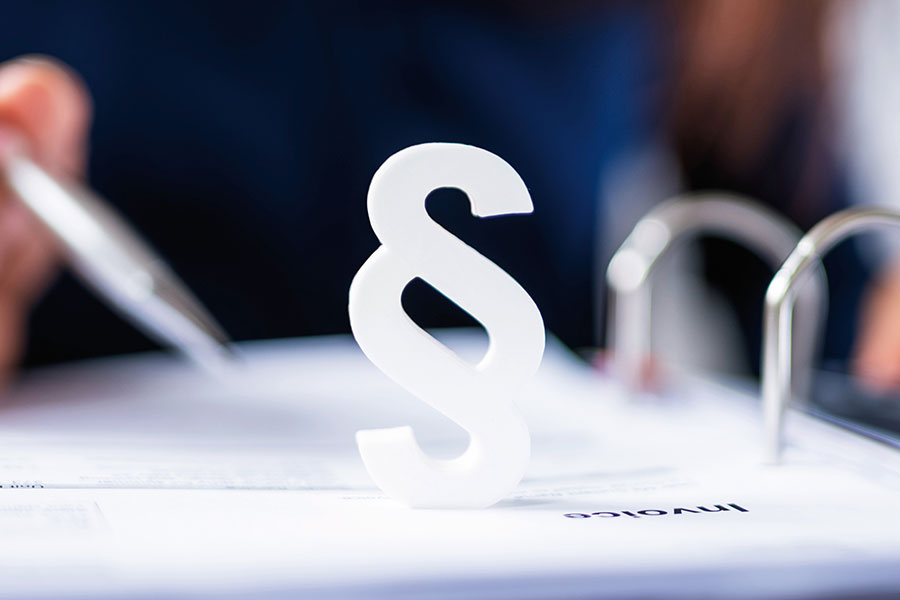 Person sitzt mit einem Kugelschreiber in der Hand vor einem Ordner, auf dem ein Paragrafenzeichen steht.