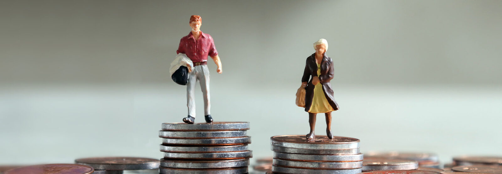 Zwei Miniatur-Figuren, ein Mann und eine Frau, stehen auf je einem Münzstapel, wobei der Münzstapel des Mannes höher ist, als der der Frau.