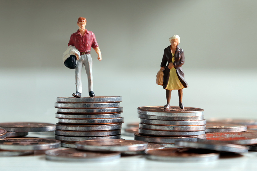 Zwei Miniatur-Figuren, ein Mann und eine Frau, stehen auf je einem Münzstapel, wobei der Münzstapel des Mannes höher ist, als der der Frau.