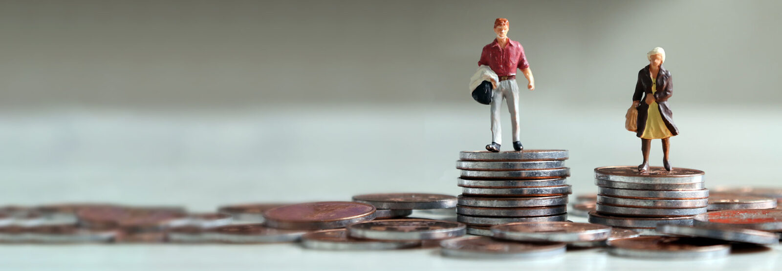 Zwei Miniatur-Figuren, ein Mann und eine Frau, stehen auf je einem Münzstapel, wobei der Münzstapel des Mannes höher ist, als der der Frau.