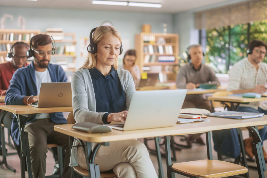 Erwachsene Männer und Frauen verschiedenen Alters sitzen an je einem Schreibtisch in einem Schulungsraum und arbeiten mit Headsets an ihren Laptops.